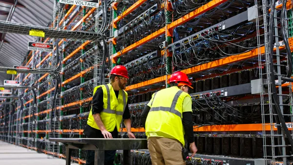 Two technicians wearing yellow vests walking past racks of electronic equipment in a data center for cryptocurrency mining.