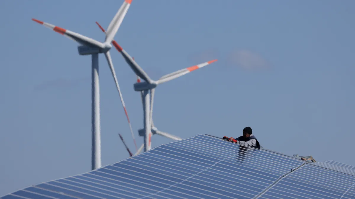 A worker installs solar panels at the construction site of a new solar energy park as wind turbines spin behind on April 06, 2023 near Prenzlau, Germany.