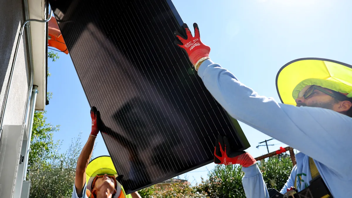 Two people wearing yellow wide-brimmed hats and orange work gloves hold a solar panel leaning on a home's roof.