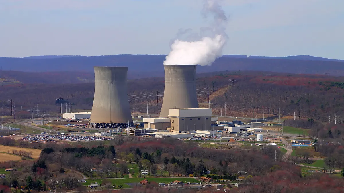 Steam rises from one of two large cement towers, with buildings and a parking lot in the foreground.