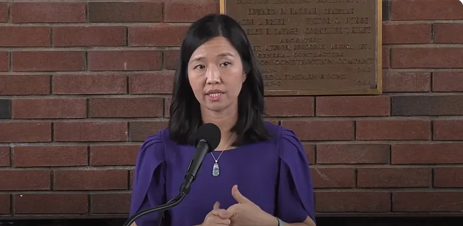 A person speaking into a microphone in front of a brick wall